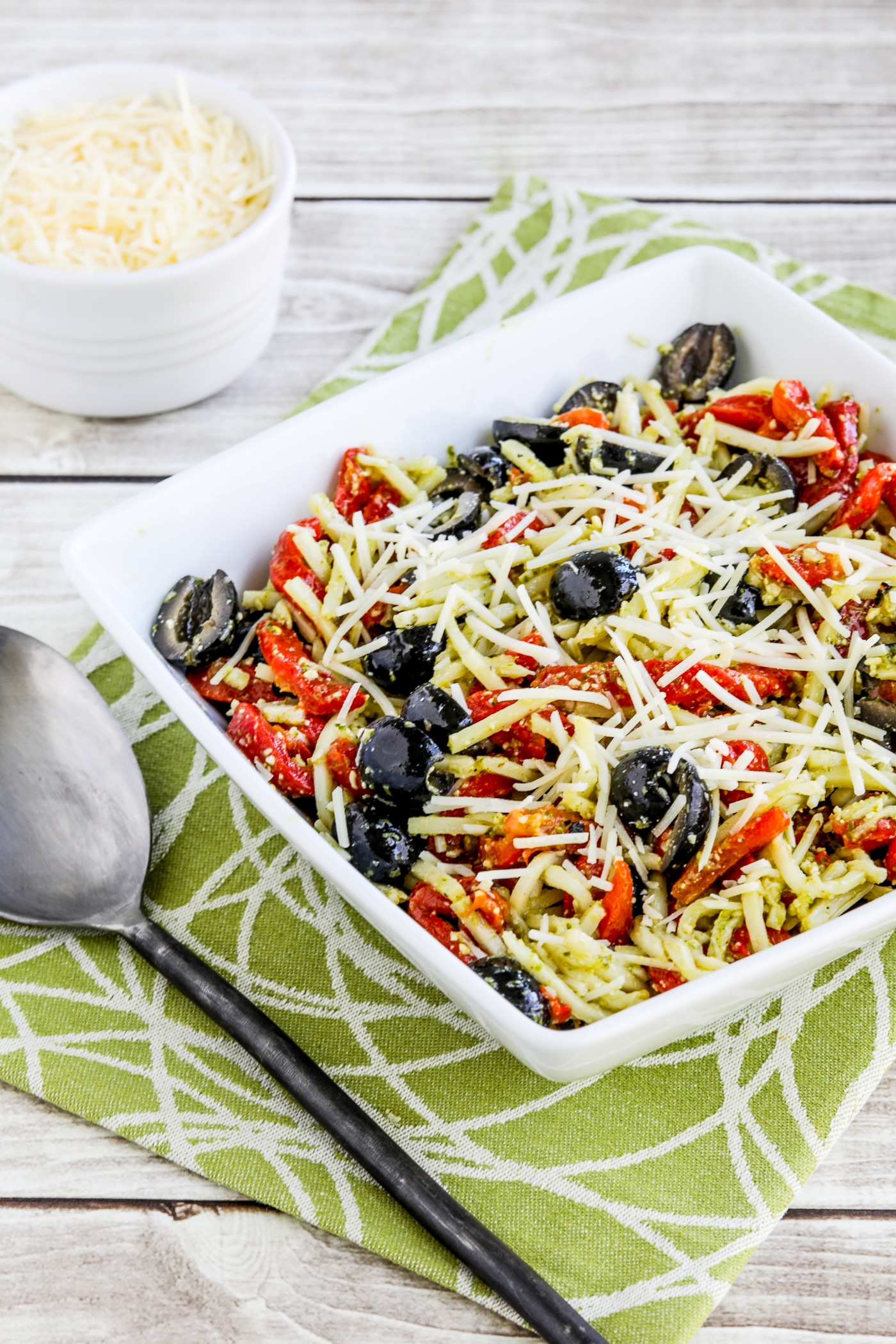 Pesta Pasta Salad shown in square serving bowl with serving spoon and Parmesan on the side.