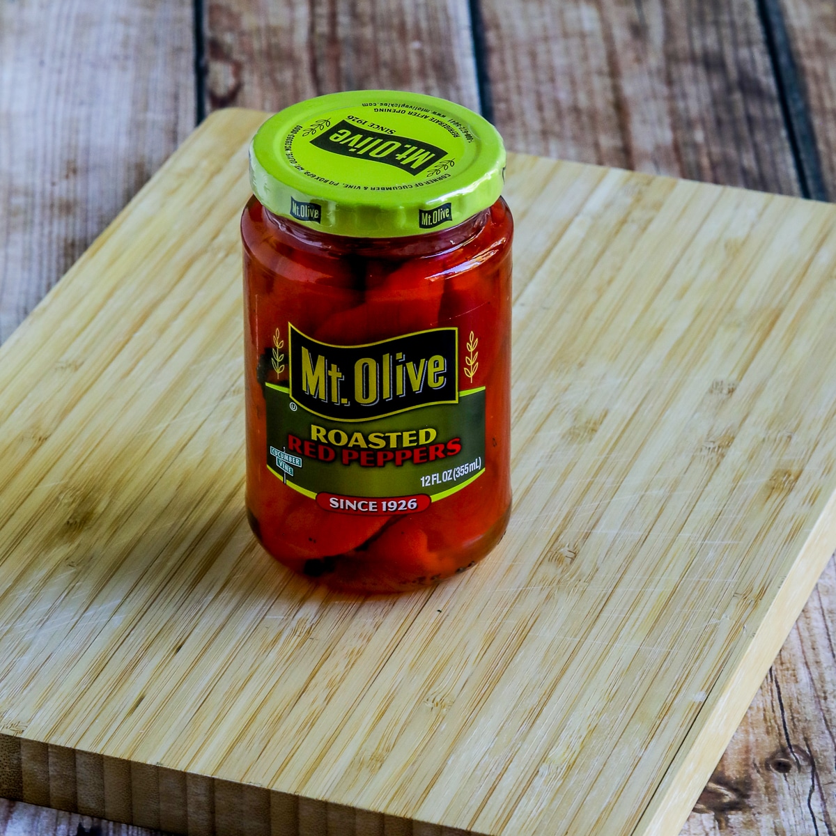 Square image of Roasted Red Peppers in a jar shown on cutting board.