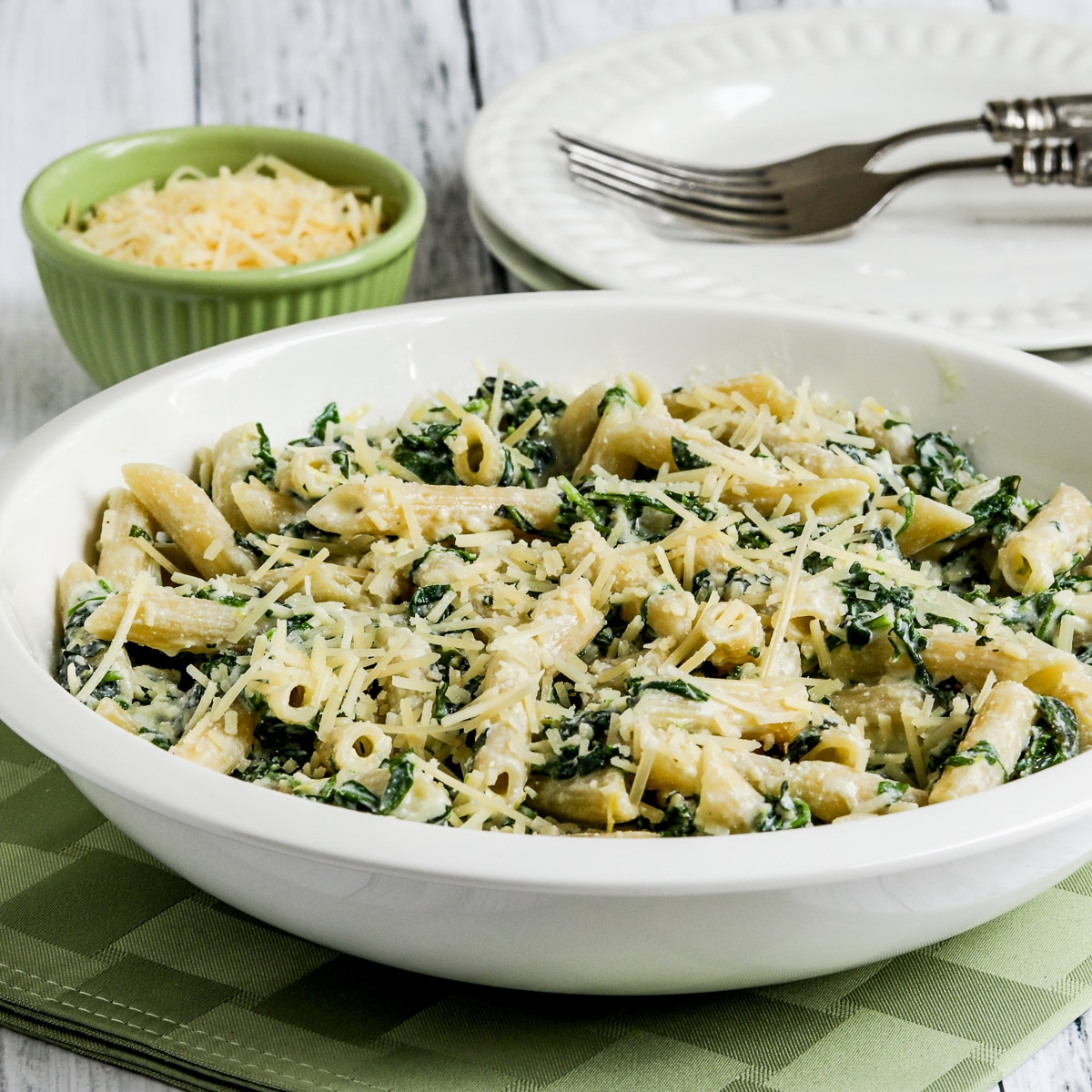 Pasta with Creamy Arugula Sauce in serving bowl with plates, forks, and extra Parmesan.