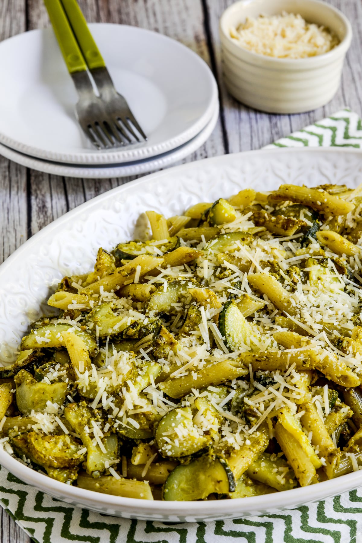 Zucchini Pesto Pasta on serving platter with plates, forks, and Parmesan
