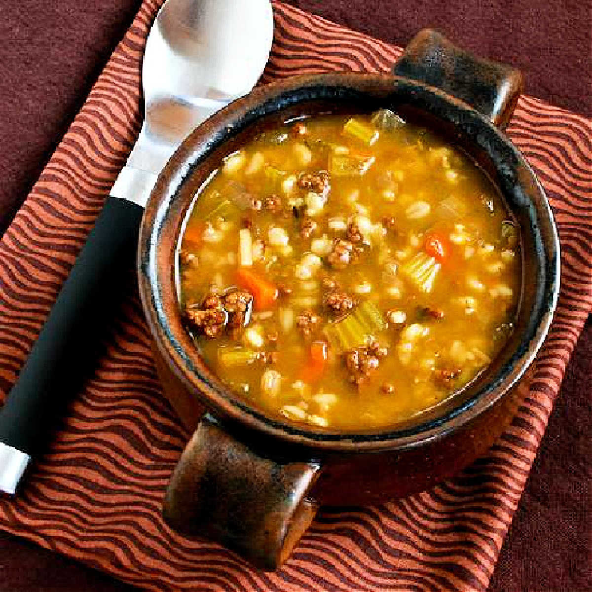 Ground Beef Barley Soup in brown bowl on wavy-striped napkin.