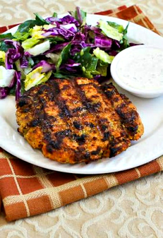 Grilled salmon burgers shown on serving plate with caper mayo and salad in background.