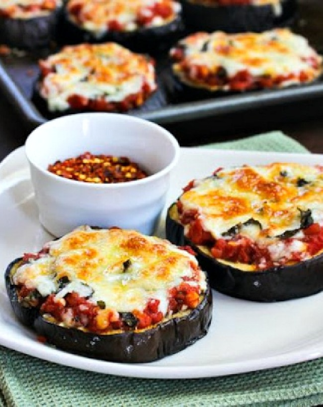 Julia Child's Eggplant Pizzas shown on serving dish with red pepper flakes and eggplant pizza on roasting pan in background