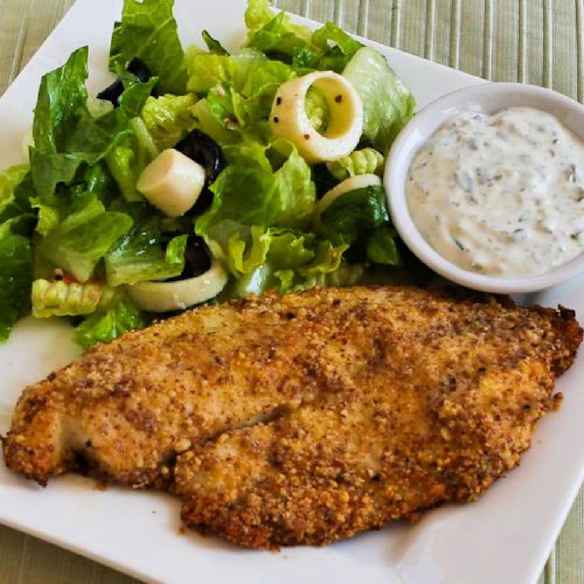 Square image of Almond and Parmesan Baked Fish shown on serving plate with salad and tartar sauce.