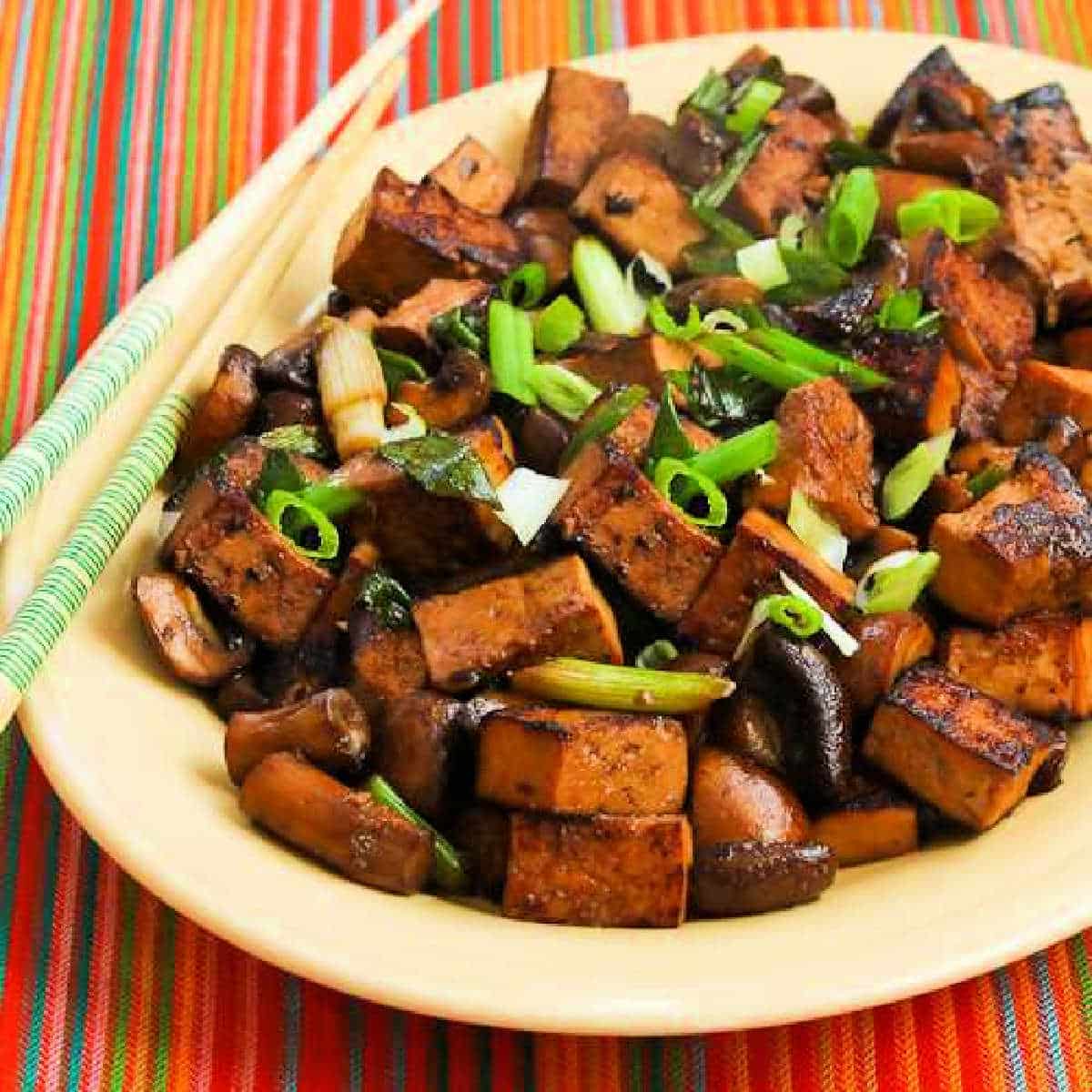 Square image of Stir-Fried Marinated Tofu with Mushrooms shown on serving plate with chopsticks.