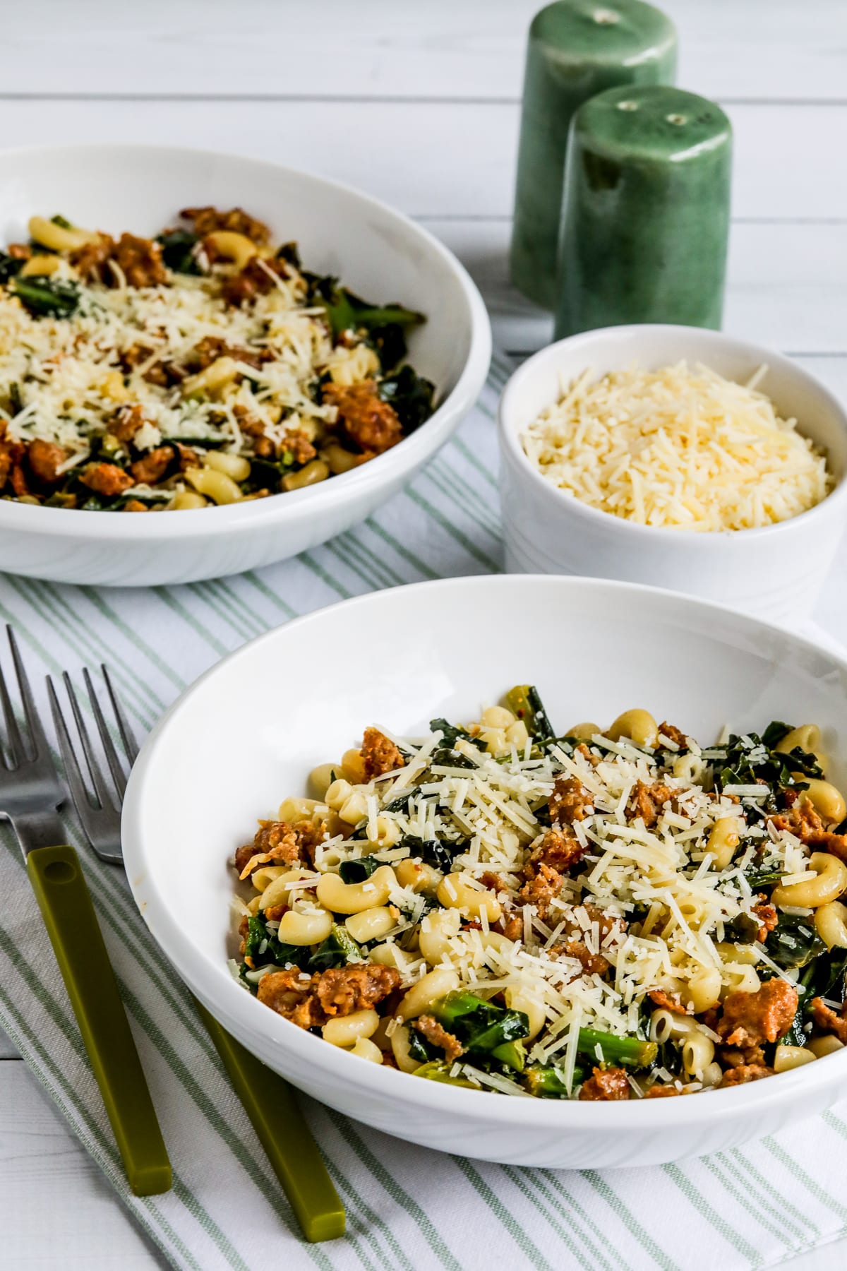 Italian Sausage Pasta with Collard Greens shown in two bowls with Parmesan and green salt and pepper shakers.