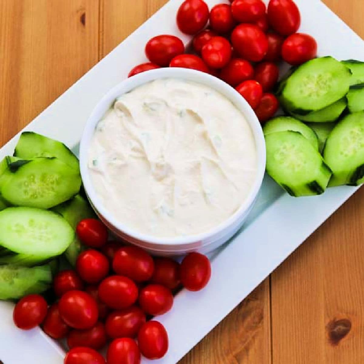 Square image of Greek Yogurt Vegetable Dip in bowl on serving platter with tomatoes and cucumbers.