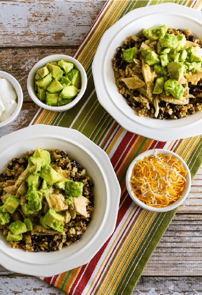 Slow Cooker Green Chile Chicken Burrito Bowl shown in two serving bowls with cheese, avocado, and sour cream on the side