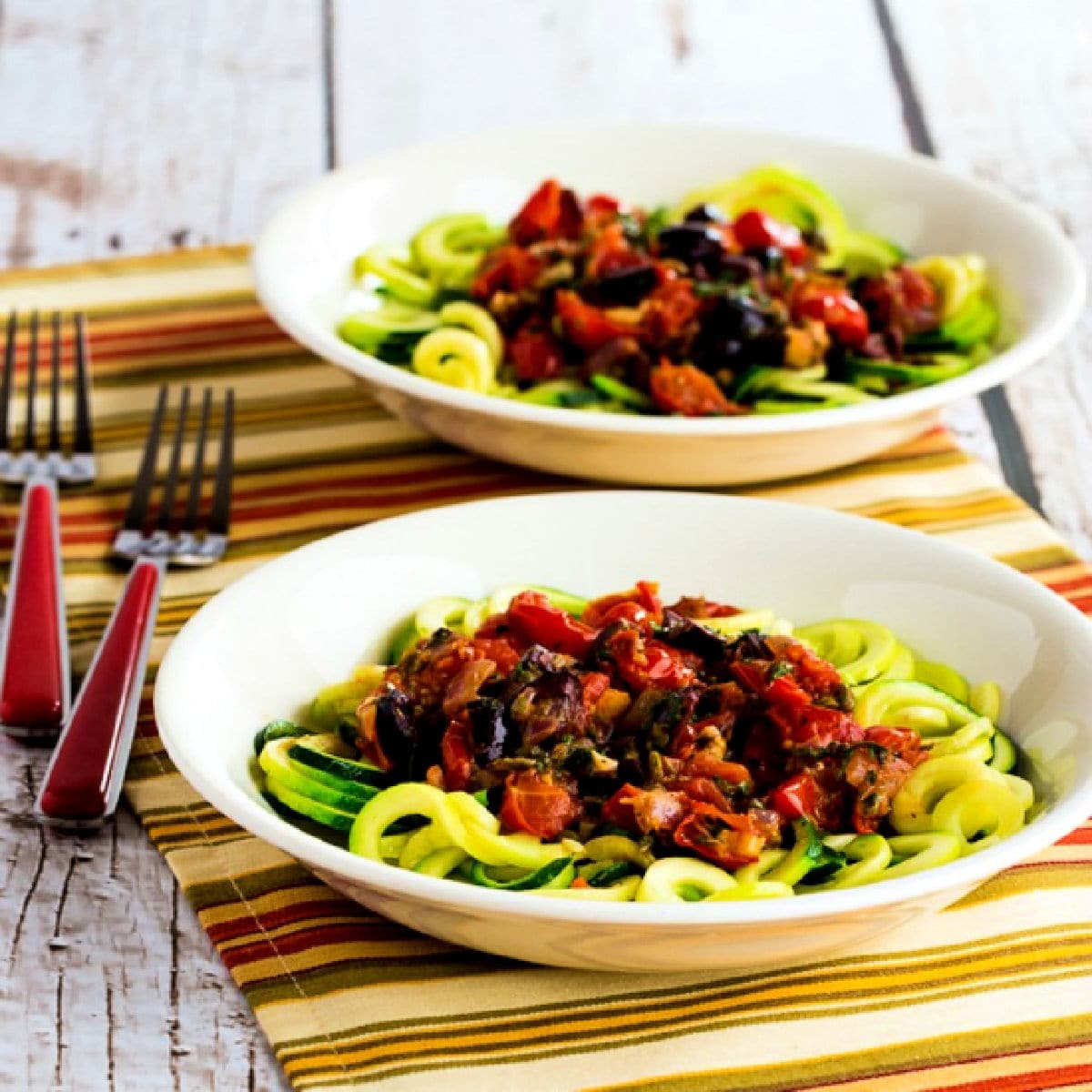 Square image for Mediterranean Zucchini Noodles shown in two bowls with forks on striped napkin.