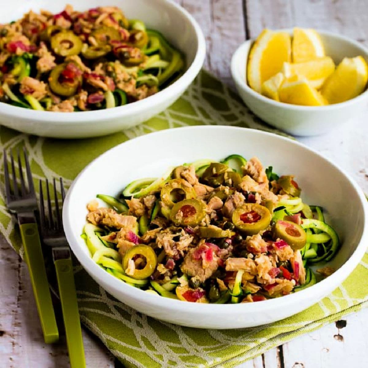 Square image of Tuna Zucchini Noodles with Green Olives shown in two serving bowls with lemons on the side.