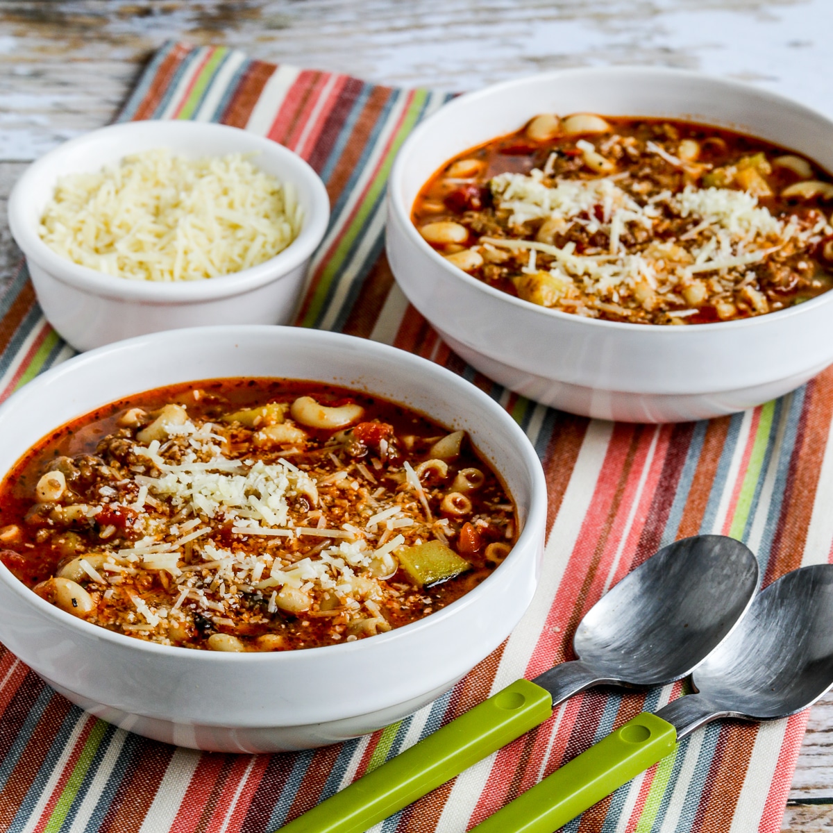 Square image of italian sausage, zucchini and macaroni soup, soup in two bowls, striped napkins, spoon and parmesan cheese.