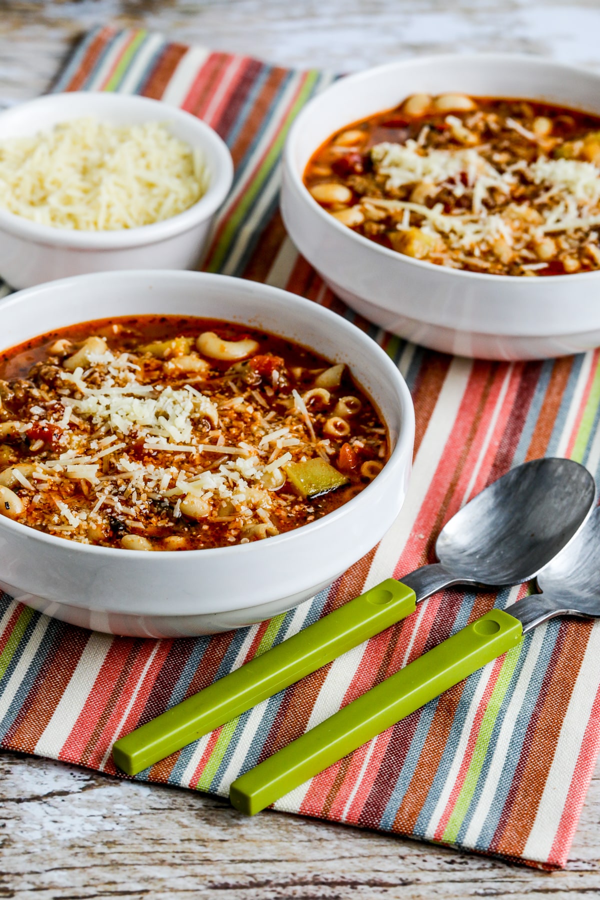 Farther back photo of Italian Sausage, Zucchini, and Macaroni Soup shown in two bowls with spoons and Parmesan