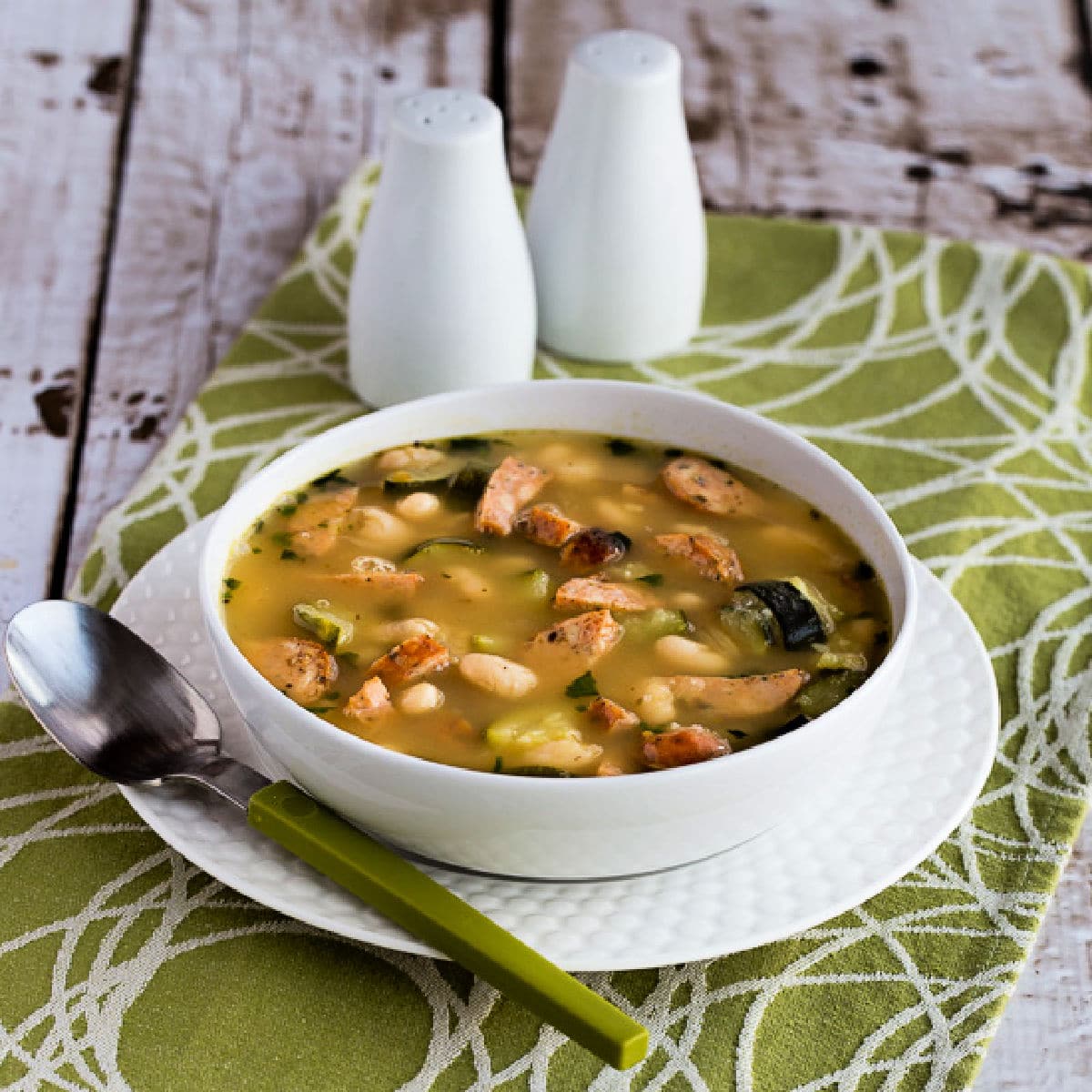 Square image of White Bean Soup with Italian Sausage, Zucchini, and Basil shown in soup bowl.
