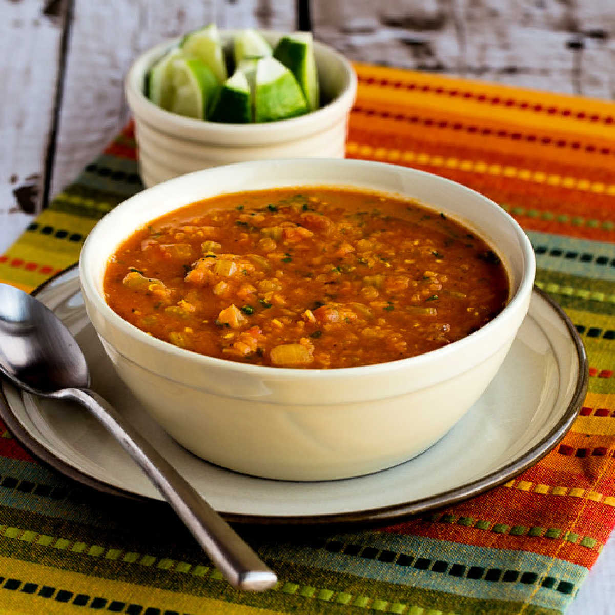 Square image of Mexican Red Lentil Stew in serving bowl on napkin, with limes in background.