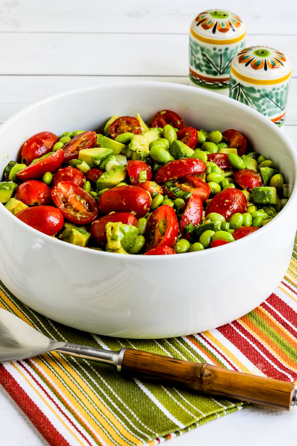 Tomato Salad with Avocado and Edamame in a Serving Bowl With Striped Napkin, Serving Fork, and Mexican Salt and Pepper Shakers.