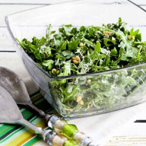 Square image of Baby Arugula Salad in glass bowl with serving forks.