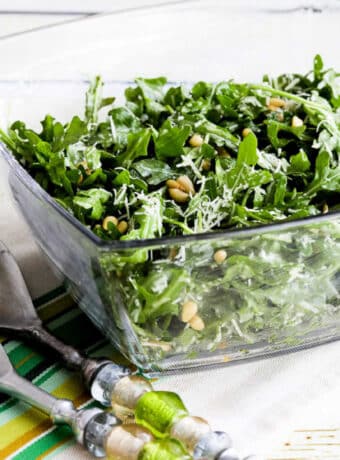 Square image of Baby Arugula Salad in glass bowl with serving forks.