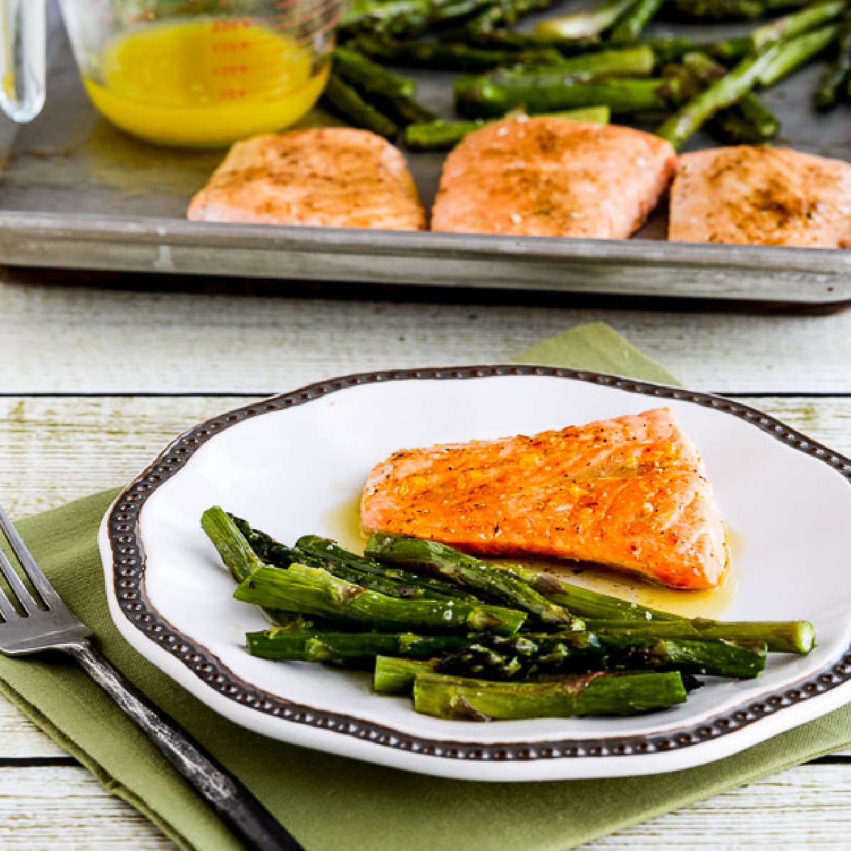 A square image of a roasted lemon salmon and asparagus sheet pan meal is presented on a plate with a sheet pan in the background.
