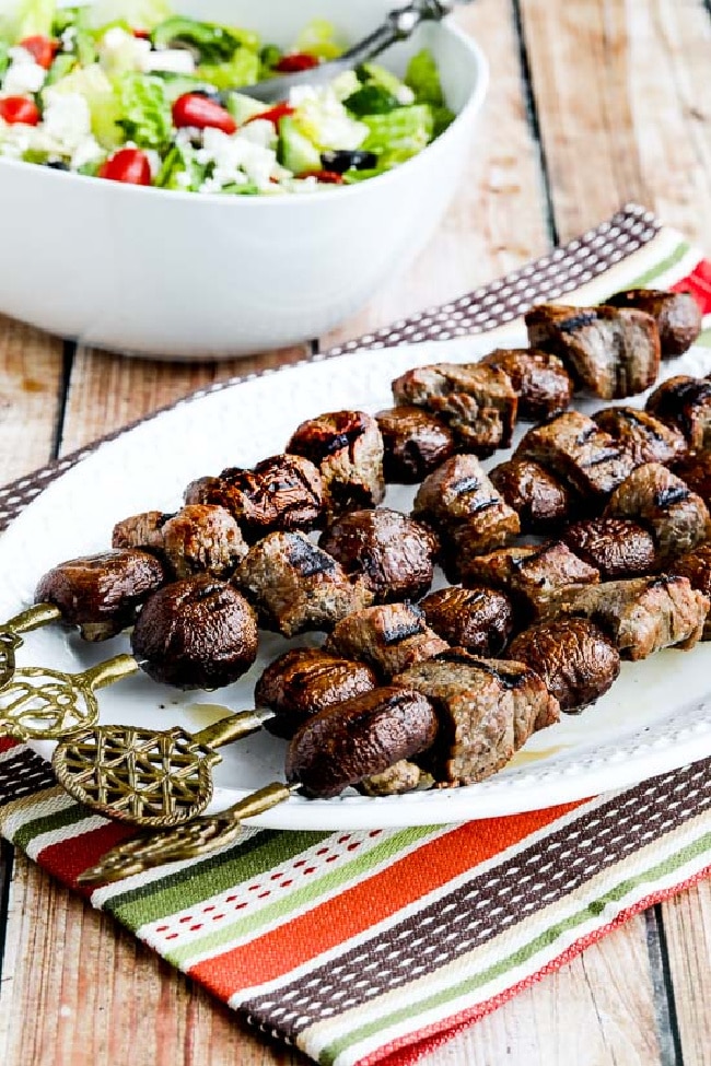 Steak and Mushroom Kabobs shown on serving plate with decorative skewers and salad in background