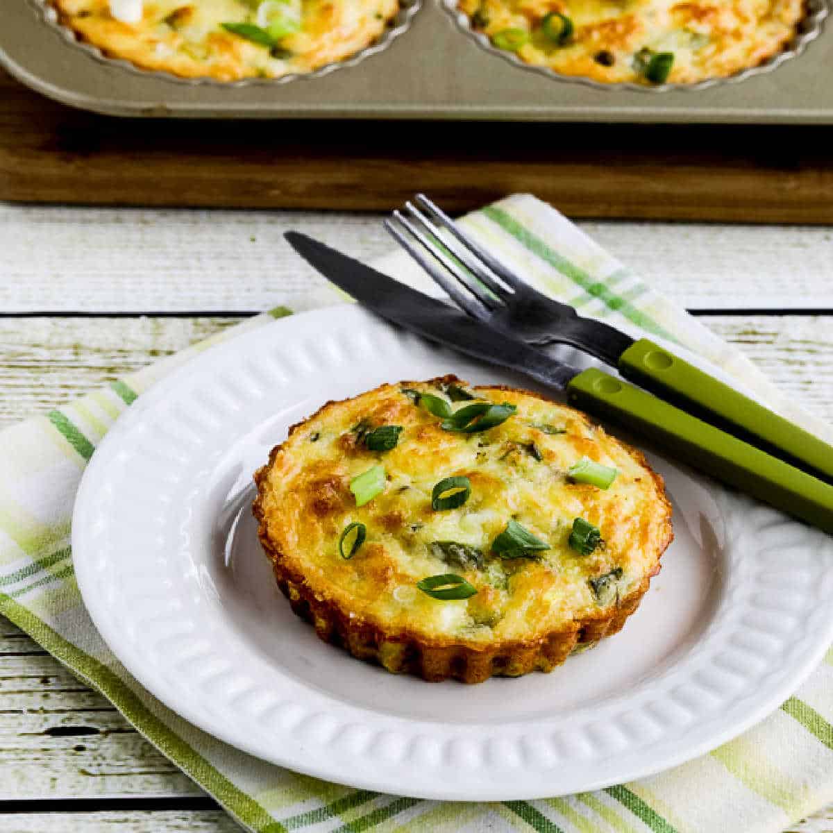 Square photo of asparagus breakfast muffins with one on a plate and a tart tray in the back