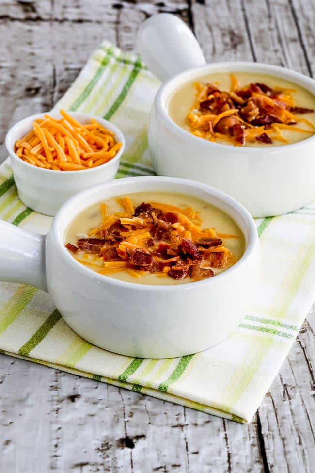 Instant cheesy cauliflower soup with bacon and green pepper clarified in two serving bowls with grated cheese on the side.