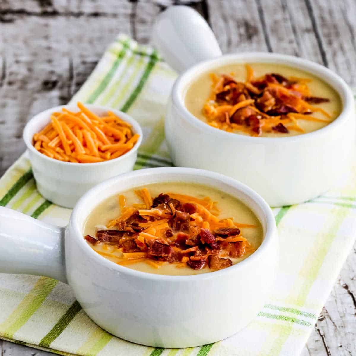 Cheesy cauliflower soup with bacon and green peppers set out in two soup bowls with cheese on the side