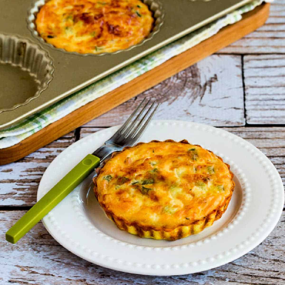 Square image for Crustless Breakfast Tarts with Asparagus shown with one breakfast tart on plate with fork, and tart pan in the back.