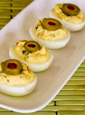 Square image of Deviled Eggs with Green Olives on serving plate on green meat.