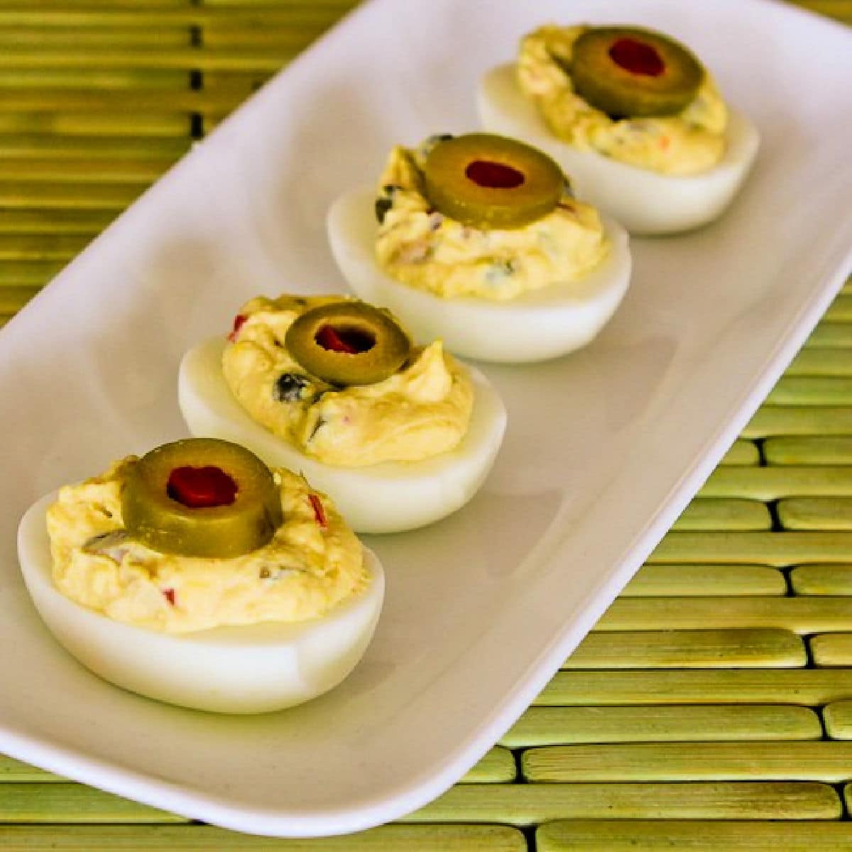 Square image of Deviled Eggs with Green Olives on serving plate on green meat.