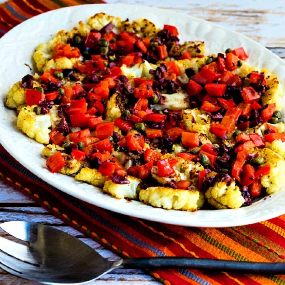 Roasted Cauliflower Steaks with Red Pepper, Capers, and Olives shown on serving platter with serving spoon
