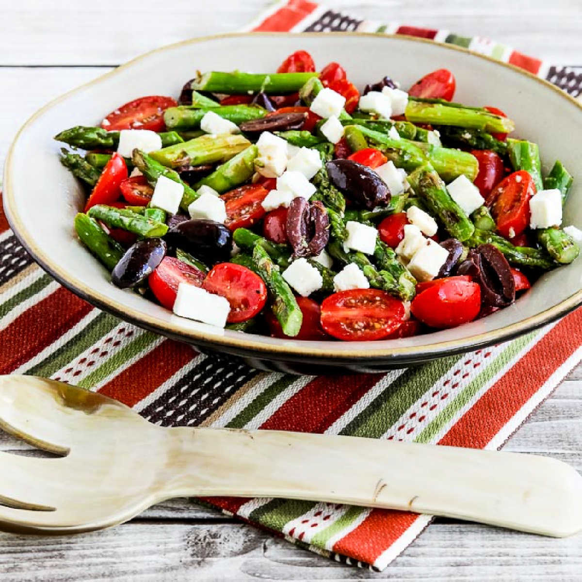 Square image of Asparagus Salad with Tomatoes, Olives, and Feta in serving bowl on colorful napkin.