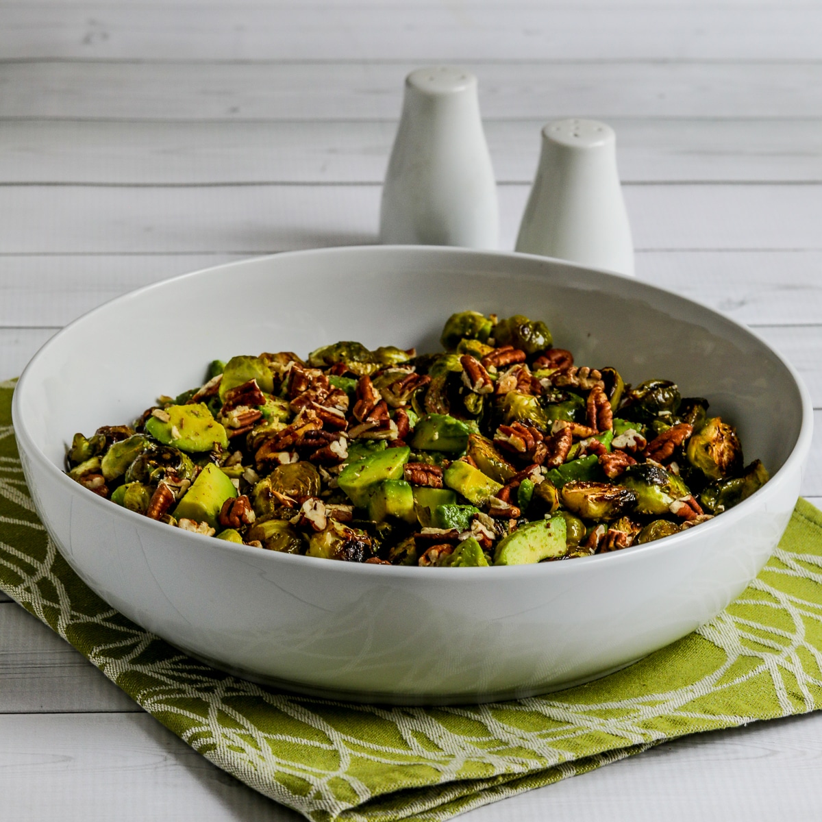 square image of Roasted Brussels Sprouts with Avocados and Pecans in serving dish with salt-pepper in background