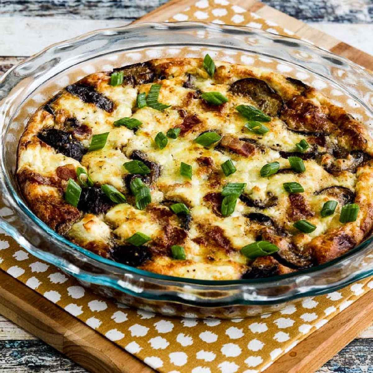 Square image of Bacon, Mushroom, and Feta Crustless Quiche shown in baking dish on napkin and cutting board