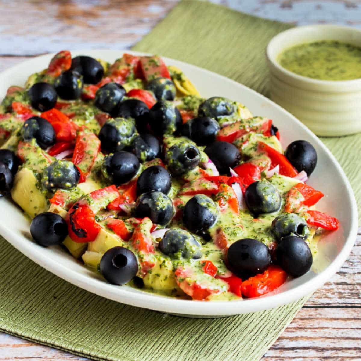 Square image of Antipasto Artichoke Salad with Basil Vinaigrette shown on serving platter on green napkin.