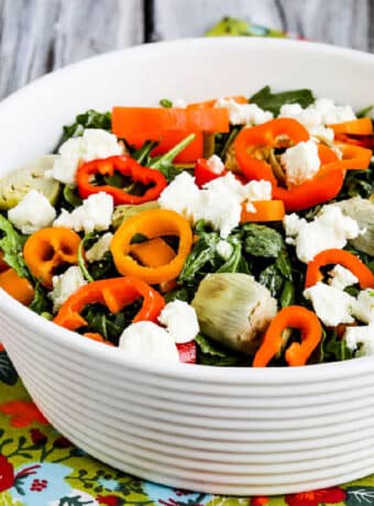 Square image of Arugula Salad with Goat Cheese, Artichokes, and Peppers shown in white bowl on colorful napkin.