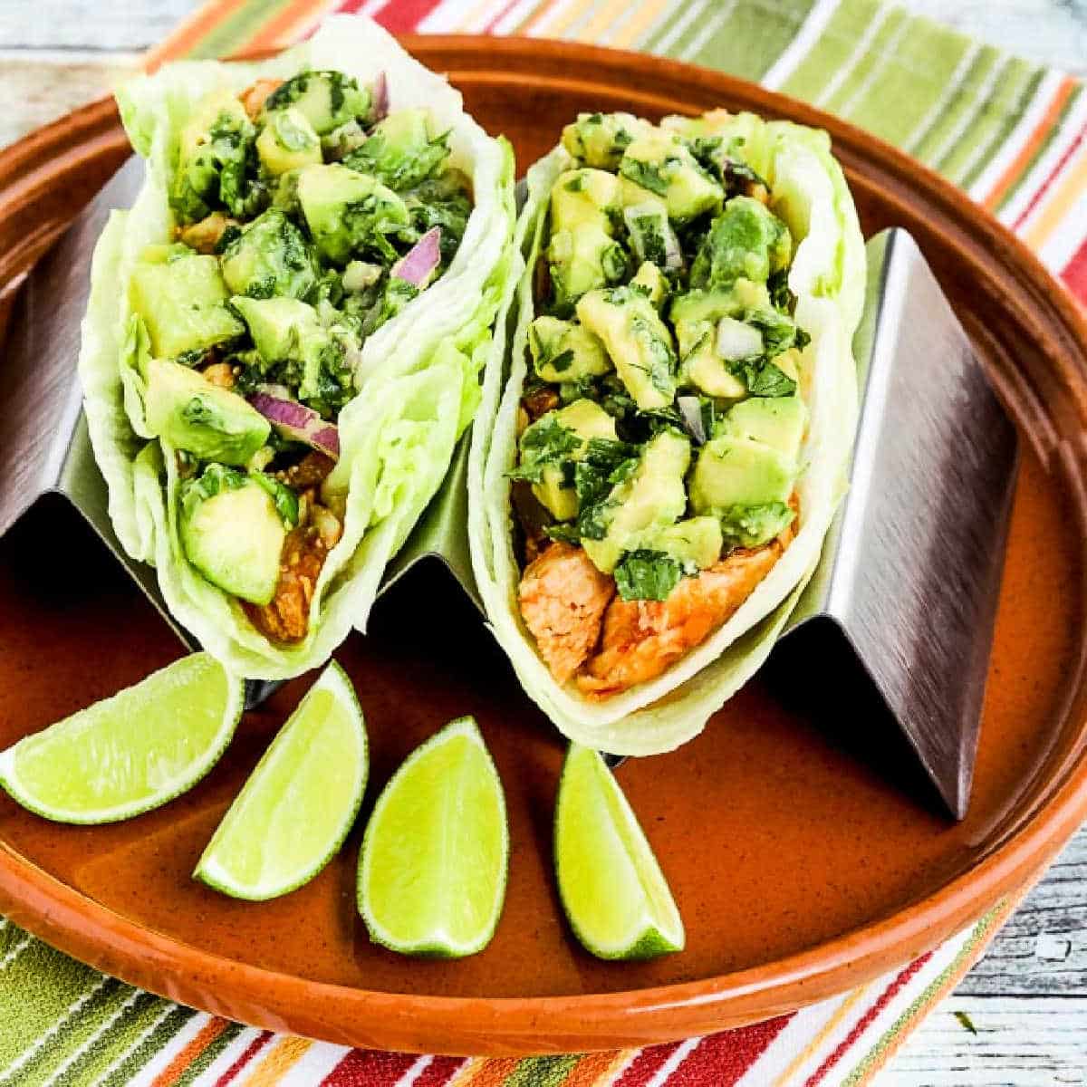 Square image of Spicy Shredded Chicken Lettuce Wrap Tacos on serving plate in taco holder, with limes.