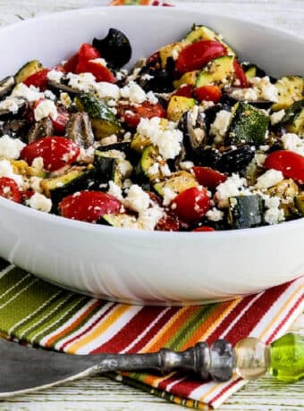 Square image of Grilled Zucchini Greek Salad shown in serving bowl on striped napkin.