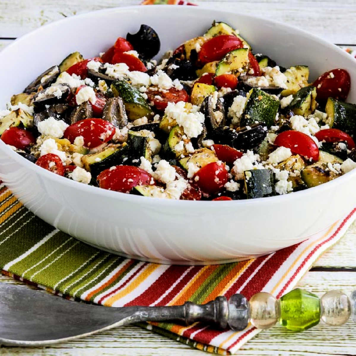 Square image of Grilled Zucchini Greek Salad shown in serving bowl on striped napkin.