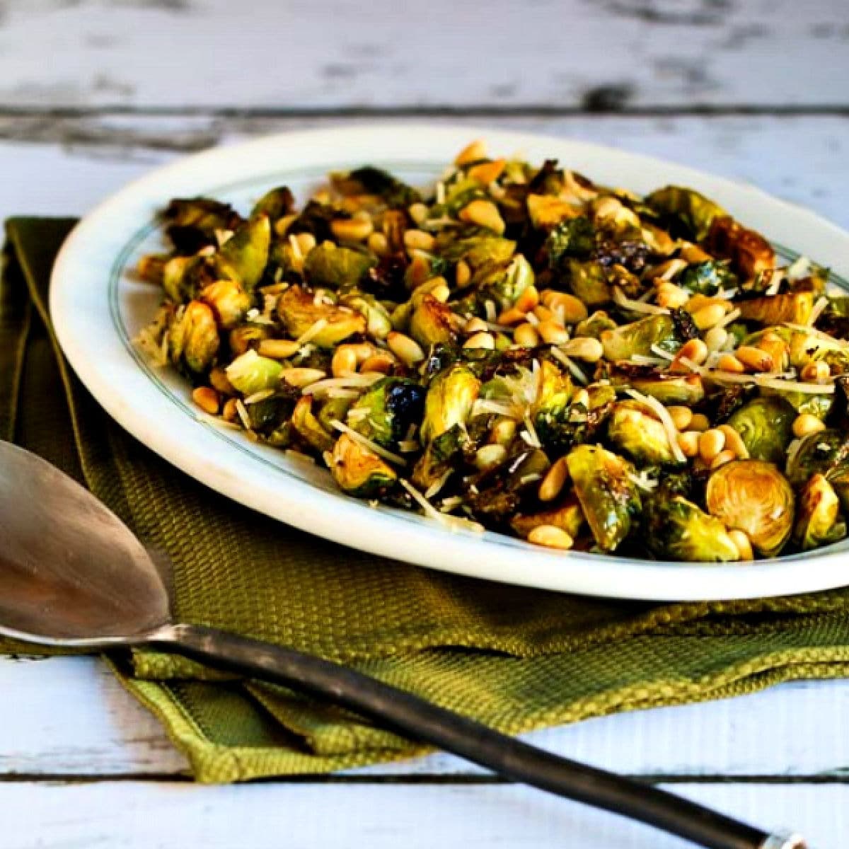 Square image for Balsamic Roasted Brussels Sprouts with Parmesan and Pine Nuts shown on serving plate with napkin and serving spoon.