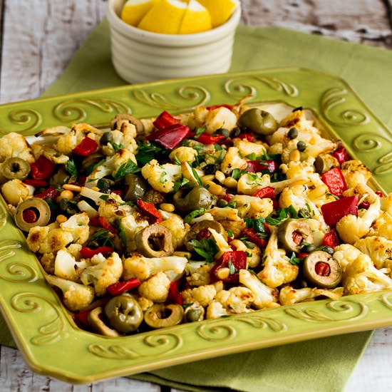 Roasted cauliflower with red peppers, green olives and pine nuts is seen on a serving plate with lemon in the background