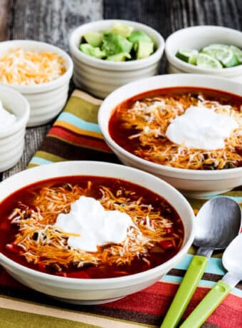 Square image of Instant Pot Taco Soup shown in two serving bowls with sour cream, cheese, limes, and avocado in background.