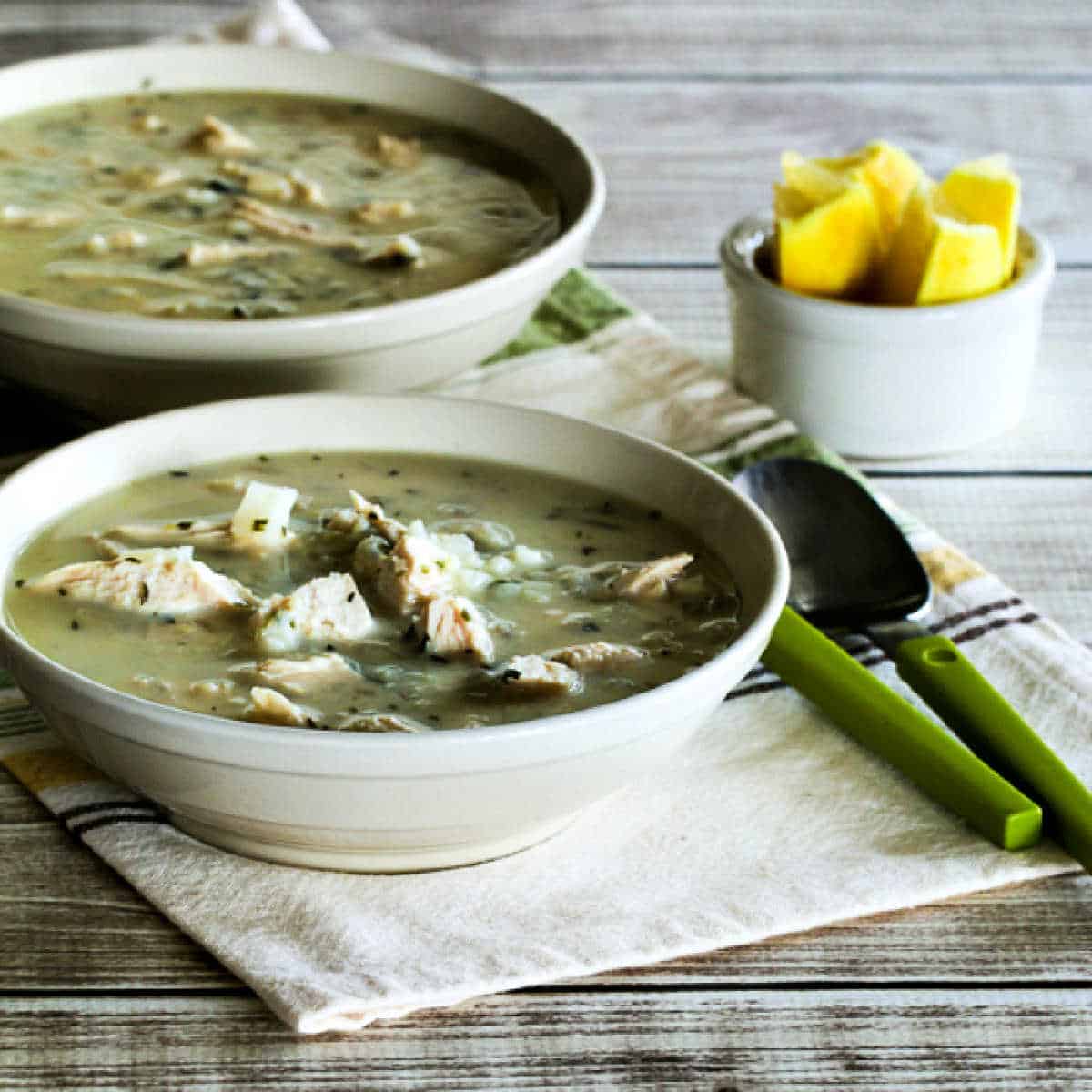 Square image of Greek lemon chicken soup in two bowls with lemon wedges on the sides.