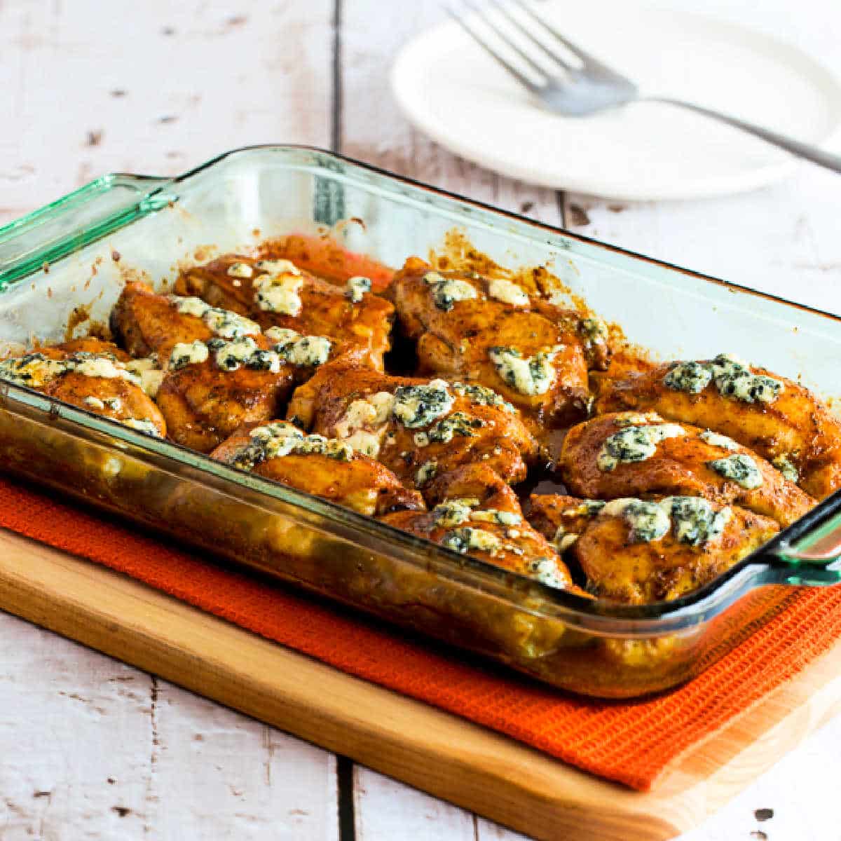 Square image of Baked Buffalo Chicken with Melted Blue Cheese shown in baking dish on cutting board.