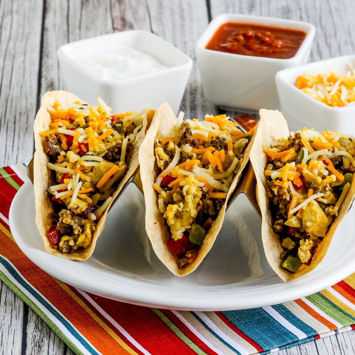 Square image of Low-Carb Breakfast Tacos Recipe with three tacos on plate and toppings in back.