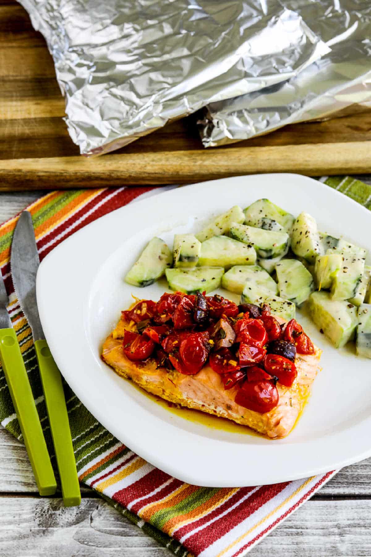 Salmon foil packet with tomatoes and olives on serving plate with pack of salmon on cutting board background