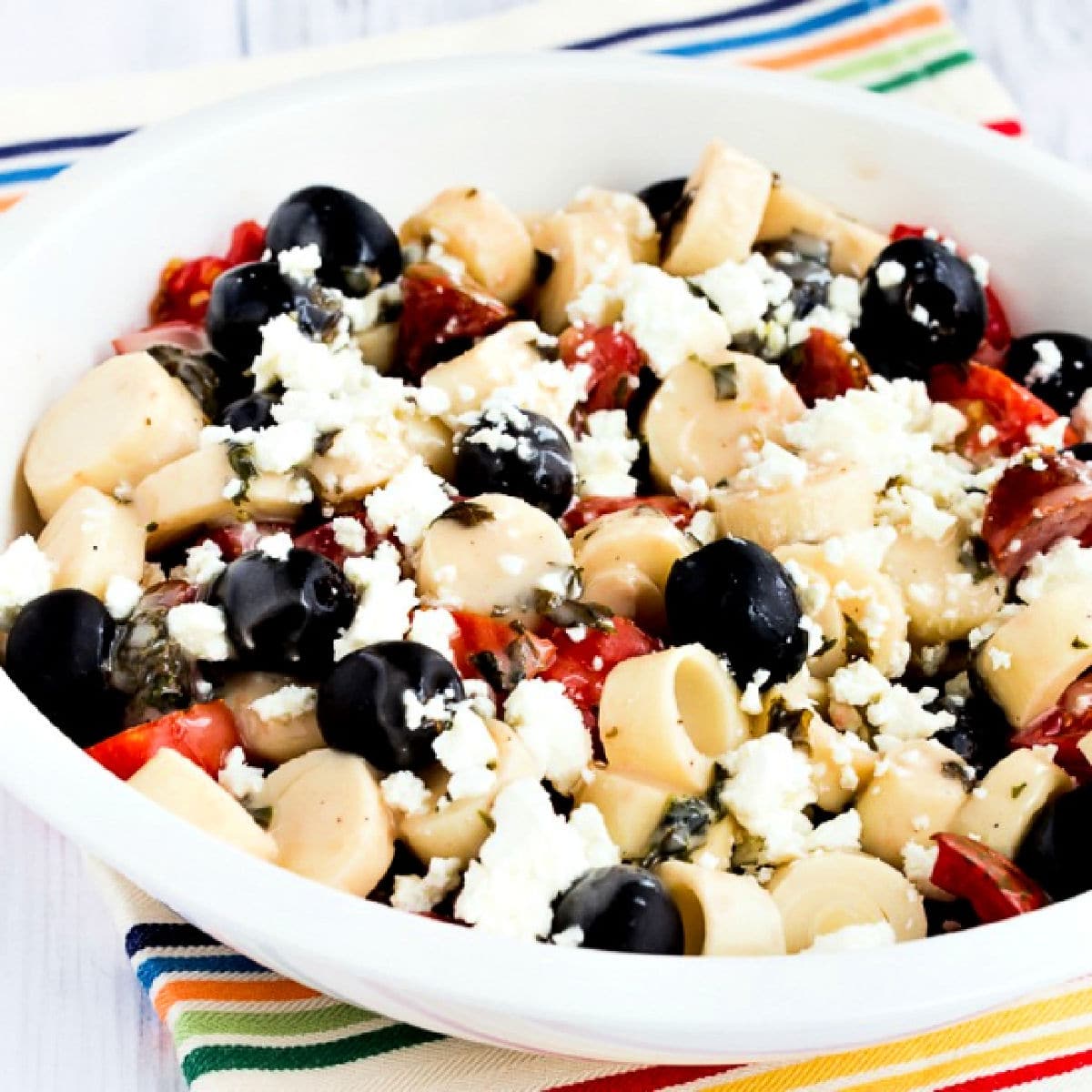Square image for Tomato Salad with Hearts of Palm, Olives, and Feta shown in white serving bowl.