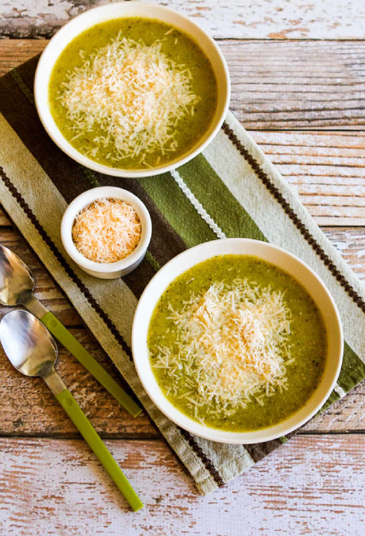 two bowls of zucchini soup on striped napkin, with green spoons and parmesan