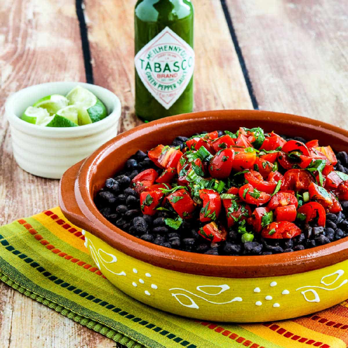 Spicy Black Beans with Cilantro shown in serving bowl with limes and Green Tabasco Sauce.