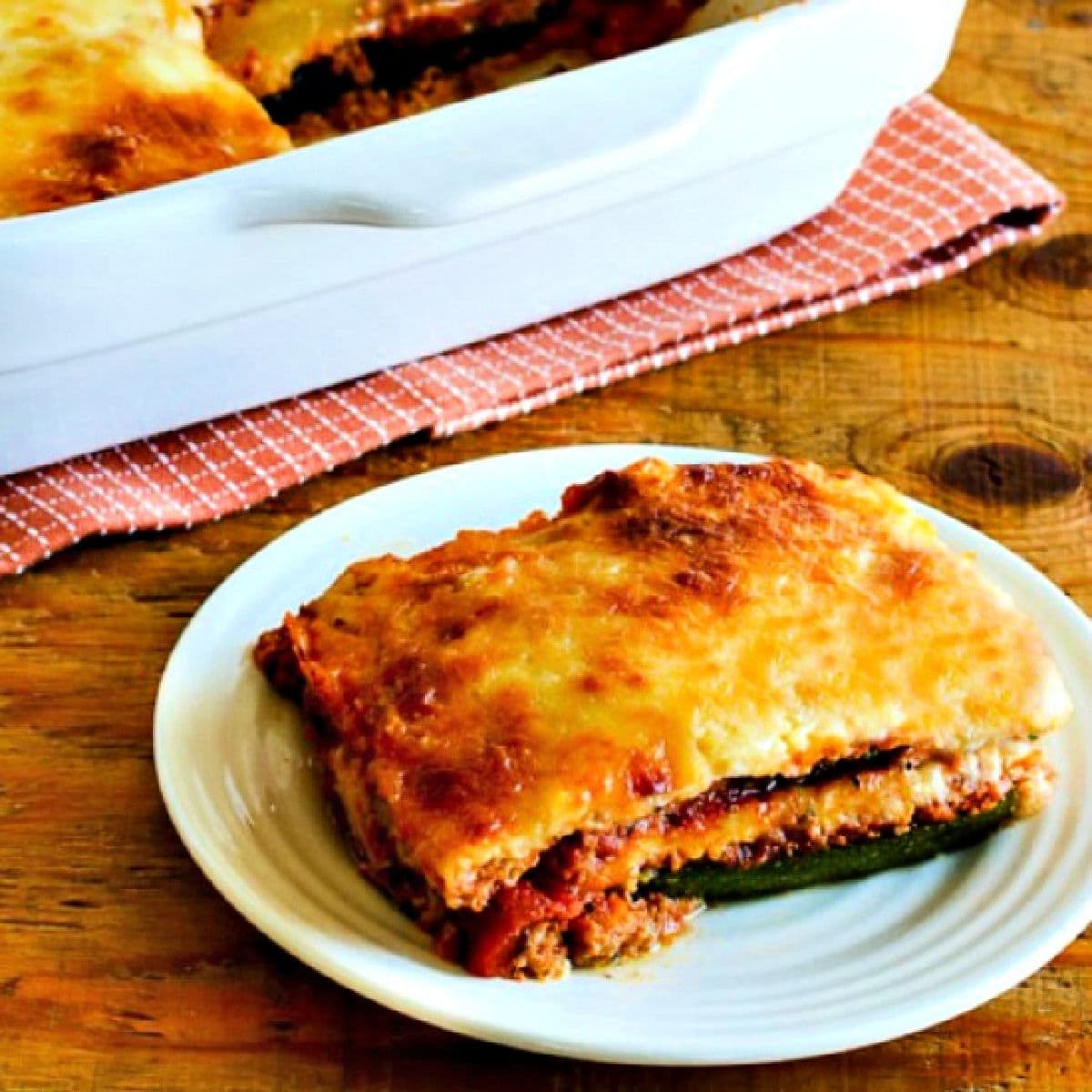 Square image of zucchini lasagna on serving plate with baking dish in back.