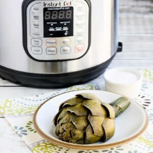 Square image of artichoke on plate and Instant Pot in back for How to Cook Artichokes in the Instant Pot